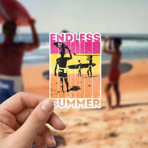 Op de afbeelding: Een sticker met een strandscène met silhouetten van surfers die surfboards dragen. De sticker heeft de woorden "ENDLESS SUMMER" in een roze en gele blok. De achtergrond toont een wazig strand en oceaan.