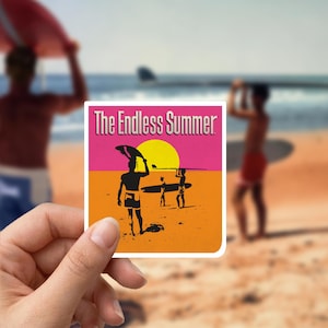 Op de afbeelding: Een sticker met de tekst "The Endless Summer" in een retro-ontwerp. De afbeelding toont silhouetten van surfers op een strand, met een gele zon en een roze en oranje achtergrond. De sticker wordt in een hand gehouden.