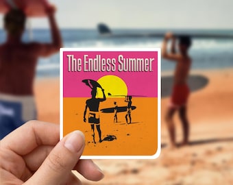 Puede incluir: Una pegatina con el texto "The Endless Summer" en un diseño retro. La imagen muestra siluetas de surfistas en una playa, con un sol amarillo y un fondo rosa y naranja. La pegatina está sujeta en una mano.