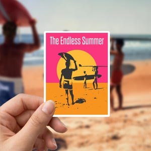 Op de afbeelding: Een rechthoekige sticker met de tekst "The Endless Summer" boven een afbeelding van surfers op een strand. De afbeelding toont silhouetten van surfers met surfplanken, een gele zon en een roze en oranje achtergrond.