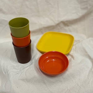 Könnte beinhalten: Ein Set aus farbenfrohem Kunststoffgeschirr, darunter drei gestapelte Becher in Grün, Orange und Braun, ein gelber quadratischer Teller und eine orangefarbene Schüssel. Die Artikel sind auf einem weißen Stoffhintergrund angeordnet.