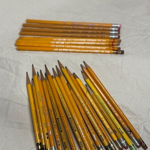 May include: A collection of yellow pencils with various brand names and numbers printed on them. The pencils have pink or green erasers and are arranged on a light-colored surface. Some of the text on the pencils includes "MONGOL" and "VELVET".