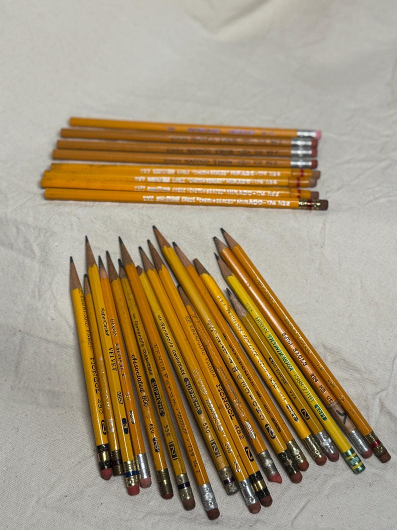 May include: A collection of yellow pencils with various brand names and numbers printed on them. The pencils have pink or green erasers and are arranged on a light-colored surface. Some of the text on the pencils includes "MONGOL" and "VELVET".