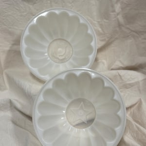 May include: Two white, fluted gelatin molds. Each mold has a scalloped edge and a star or floral design in the center. The molds are made of a translucent, off-white plastic and are likely vintage kitchenware.