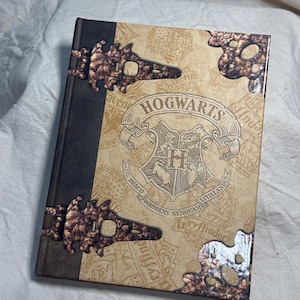 May include: A hardcover book with a tan cover featuring the Hogwarts crest and the text "HOGWARTS" and "DRACO DORMIENS NUNQUAM TITILLANDUS". The book has a dark brown spine and decorative metallic corner accents with a textured, bronze-colored finish.