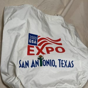 May include: White tote bag with the text "2003 GSA EXPO SAN ANTONIO, TEXAS" in red and blue. The design includes a stylized American flag and a cactus wearing a cowboy hat.