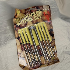 Peut inclure: Un ensemble vintage de casse-noix et de cure-noix dans son emballage d'origine. L'emballage présente un design marron et jaune avec les mots "Nutcracker Nutpick Set". L'ensemble comprend un casse-noix argenté et plusieurs cure-noix argentés.