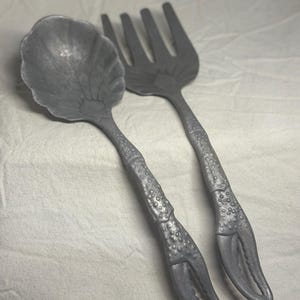 May include: A pair of metallic gray serving utensils consisting of a spoon and a fork. The handles are textured to resemble crustacean legs, ending in pincer-shaped tips. The spoon bowl is shaped like a shell, and the fork has four tines.