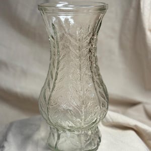 May include: A clear glass vase featuring a textured surface with a repeating embossed leaf pattern. The vase has a flared rim, a curved body that tapers toward the base, and a wide, circular foot. The glass is transparent with a subtle sheen.
