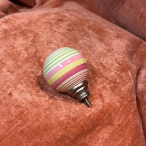 May include: A spherical cabinet knob featuring horizontal stripes in shades of pink, yellow, and light green. The knob has a smooth, glossy finish and is attached to a metallic, silver-toned base with a threaded screw for installation.