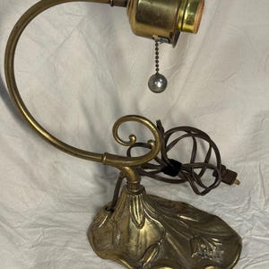 Peut inclure: Une base de lampe de table en laiton avec une base sculptée en forme de feuille et un bras incurvé. La lampe comprend une douille cylindrique avec une chaînette et une petite tirette métallique sphérique. Un cordon électrique marron est fixé.