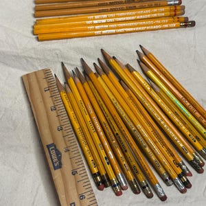 May include: A collection of yellow pencils with erasers and text, alongside a wooden ruler. The pencils are arranged on a light-colored surface. The pencils have various brand names printed on them, including "EAGLE" and "MONGOL".