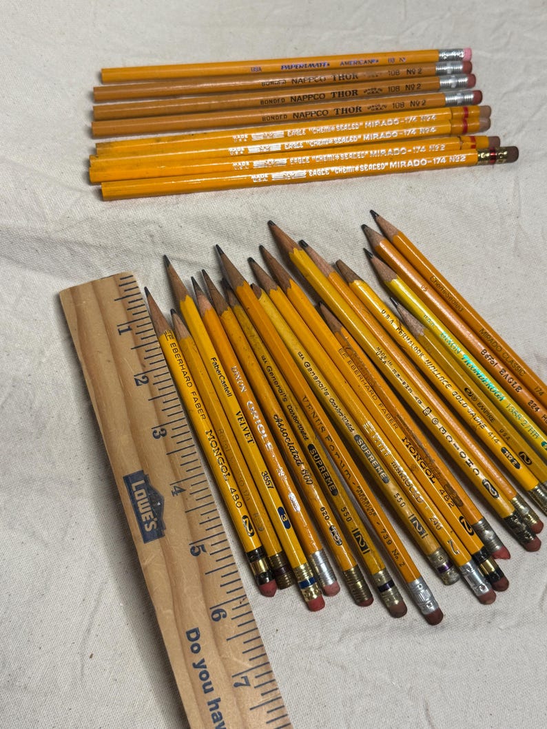 May include: A collection of yellow pencils with erasers and text, alongside a wooden ruler. The pencils are arranged on a light-colored surface. The pencils have various brand names printed on them, including "EAGLE" and "MONGOL".