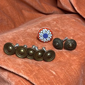 May include: A collection of hardware knobs including five circular antique brass-finished knobs with a concentric ring design, two ornate dark metal knobs with embossed detailing, and one round ceramic knob featuring a blue and white flower pattern.