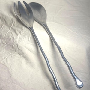 May include: A pair of silver-toned metal salad servers consisting of a three-pronged fork and a deep-bowled spoon. Both utensils feature long, slender, and irregularly wavy handles that terminate in rounded, bulbous ends with a smooth finish.