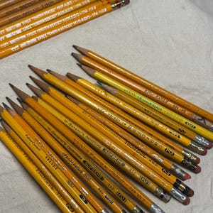 May include: A collection of yellow pencils with various brand names and model numbers printed on them. The pencils have silver ferrules and pink or red erasers. Some pencils are labeled "EAGLE MIRADO" and "MONGOL".