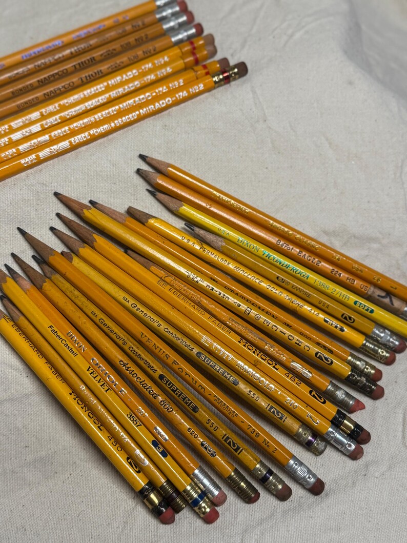 May include: A collection of yellow pencils with various brand names and model numbers printed on them. The pencils have silver ferrules and pink or red erasers. Some pencils are labeled "EAGLE MIRADO" and "MONGOL".