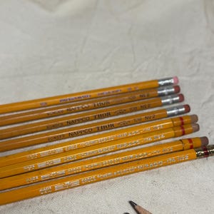 May include: A collection of yellow pencils with silver lettering and pink erasers. The pencils are labeled with the brand names "NAPPCO THOR" and "EAGLE MIRADO". The pencils are arranged on a light-colored fabric surface.