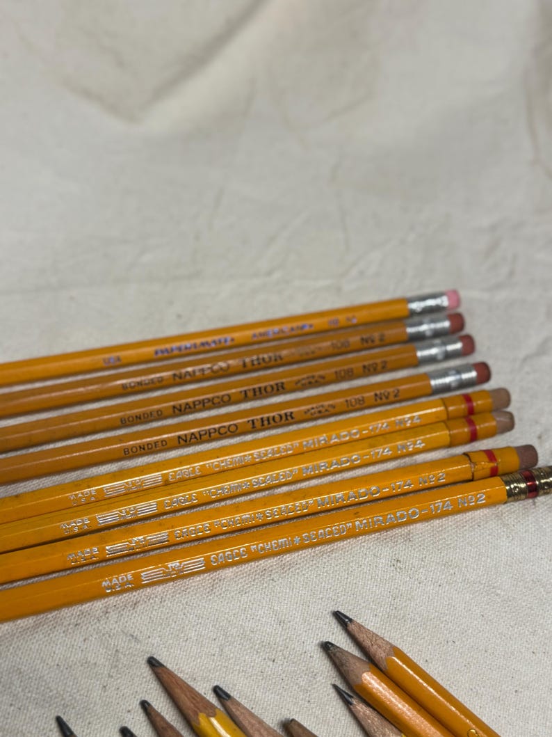 May include: A collection of yellow pencils with silver lettering and pink erasers. The pencils are labeled with the brand names "NAPPCO THOR" and "EAGLE MIRADO". The pencils are arranged on a light-colored fabric surface.