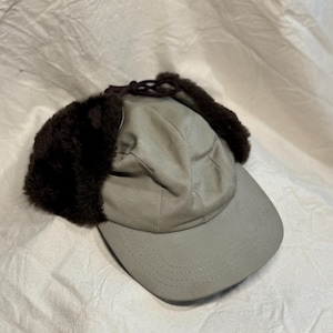 May include: A tan baseball cap with dark brown faux fur ear flaps. The cap has a curved brim and a brown drawstring closure. The hat is made of a soft, durable fabric, suitable for cold weather.