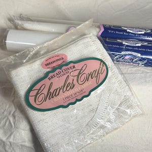May include: A Charles Craft 14 count cross stitch bread cover fabric measuring 18 inches by 18 inches, packaged in a clear plastic sleeve with a pink and green label. The fabric is off-white with a visible weave and a fringed edge detail.