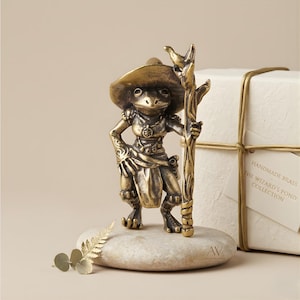 May include: A brass frog figurine, posed with a staff and hat, stands on a stone. The figurine is part of "The Wizard's Pond Collection." A gift box tied with gold string is in the background. The figurine is detailed with intricate clothing and accessories.