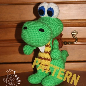 Könnte beinhalten: Grünes Häkel-Dinosaurierspielzeug mit gelben und braunen Streifen auf der Brust. Der Dinosaurier hat große weiße Augen mit blauen Pupillen und einen kleinen braunen Rucksack.