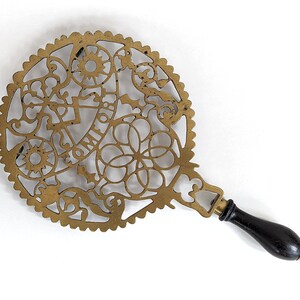 May include: A brass trivet with a decorative, intricate design and a black wooden handle. The trivet is round and has a scalloped edge. The design features a variety of geometric shapes and floral motifs.