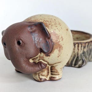 May include: A ceramic elephant planter with a brown head and ears, and a beige body with brown speckles. The planter has a separate, tree-stump-shaped container attached to the back. The elephant's trunk is curled, and it has small, black eyes.