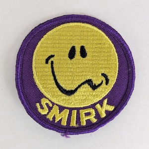 May include: Circular embroidered patch featuring a yellow smiley face with the word "SMIRK" in yellow text on a purple background. The patch is designed to be sewn or ironed onto clothing or fabric.