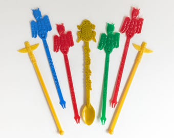 Op de afbeelding: Een verzameling kleurrijke plastic cocktailroerstaafjes. De roerstaafjes hebben verschillende ontwerpen, waaronder totempalen, vogels en een kikker. De kleuren zijn geel, blauw, rood, groen en goud. Sommige roerstaafjes hebben de tekst "COPPER ROOM".