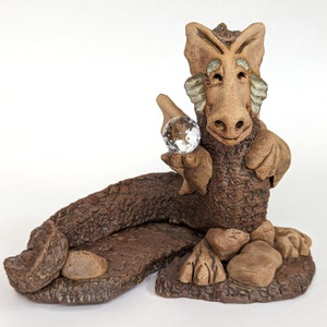 May include: A brown ceramic dragon figurine holding a clear crystal ball. The dragon is sitting on a rocky base.