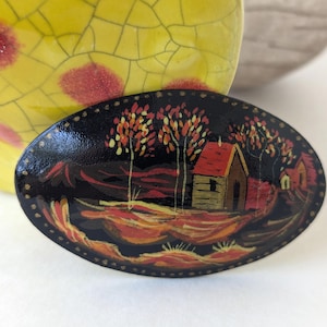Peut inclure: Broche ovale laquée noire avec une scène de paysage peinte à la main. L'œuvre représente une petite maison au toit rouge, des arbres aux feuilles orange et jaunes, et un paysage aux tons chauds. La broche a une bordure dorée.
