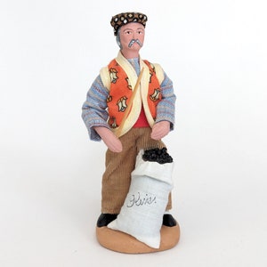May include: A figurine of a man wearing a patterned vest, a hat, and brown pants. He is holding a sack of coal that says "Heiz."