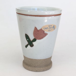 May include: A white ceramic tumbler with a brown rim and base. The tumbler features a hand-painted design of a pink flower with green leaves. A sticker with Japanese text is affixed to the side.