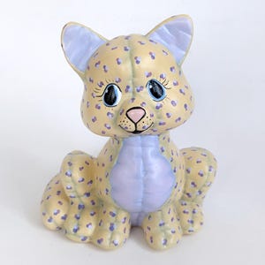 May include: A ceramic figurine of a cat with a yellow and purple spotted pattern. The cat has large blue eyes and a pink nose.