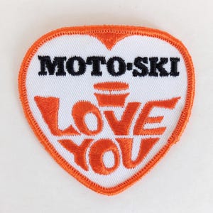 May include: Heart-shaped patch with an orange border and white center. The patch features the words "MOTO-SKI" in black and "LOVE YOU" in orange. The design includes an orange graphic above the words "LOVE YOU".
