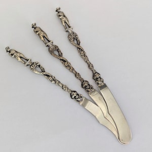 May include: Three silver butter knives with ornate handles featuring a decorative design of flowers and figures. The blades are plain and silver.