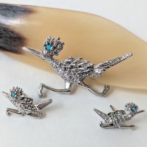 Puede incluir: Tres broches plateados con forma de correcaminos con detalles turquesa. El broche más grande está en posición de carrera, mientras que los broches más pequeños tienen un diseño similar. Los broches están colocados sobre un fondo claro.