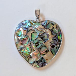 Peut inclure: Pendentif en forme de cœur avec une bélière argentée. Le pendentif est incrusté de morceaux de coquillage d'ormeau irisés, disposés en damier. La coquille affiche une gamme de couleurs, dont le vert, le bleu et le rose.
