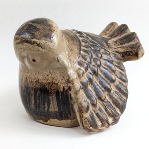 May include: A ceramic bird figurine with brown and tan glaze. The bird is perched with its wings spread out. The bird's head is turned to the side and its eyes are closed.