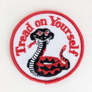 May include: Circular embroidered patch with a red border. The patch features a black and red snake with red eyes on a white background, and the words "Tread on Yourself" in red.
