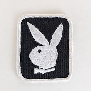 Peut inclure: Écusson brodé avec le logo de la tête de lapin blanc sur fond noir. Le patch a une bordure blanche et une forme carrée aux coins arrondis. Le lapin porte un nœud papillon.