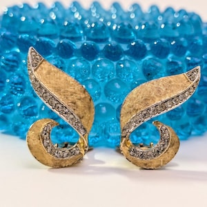 May include: A pair of gold-toned leaf-shaped clip-on earrings. Each earring features a textured gold surface with a curved, embellished edge set with small, clear crystals. The earrings are set against a blurred blue background.