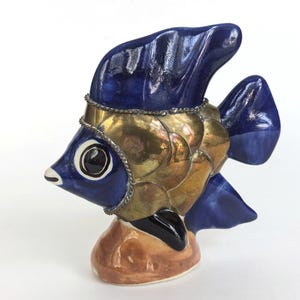 May include: A ceramic fish ornament with a blue head and fins, and gold-coloured scales. The fish has a decorative silver-coloured trim around its head and a brown base. The fish's eye is black and white.