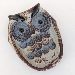 May include: A ceramic owl-shaped dish with a textured surface. The owl has large blue eyes with black centers, brown and blue feathers, and brown accents. The dish is approximately 15 cm long and 10 cm wide.