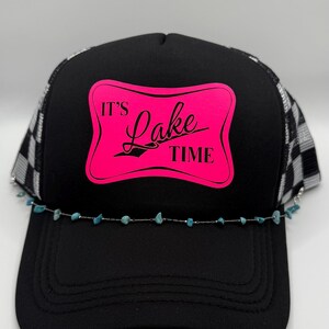 May include: Black trucker hat with a mesh back and a curved brim. The front panel features a pink rectangle with the words "It's Lake Time" in a script font. A turquoise beaded chain is attached to the brim.