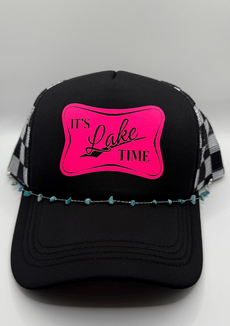 May include: Black trucker hat with a mesh back and a curved brim. The front panel features a pink rectangle with the words "It's Lake Time" in a script font. A turquoise beaded chain is attached to the brim.