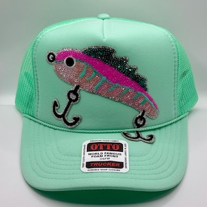 May include: A mint green trucker hat with a sequined fish design in pink, teal, and black. The hat features a curved brim and a mesh back. The hat has the text "OTTO WORLD FAMOUS FOAM FRONT OSFM TRUCKER DURABLE SNAP CLOSURE".