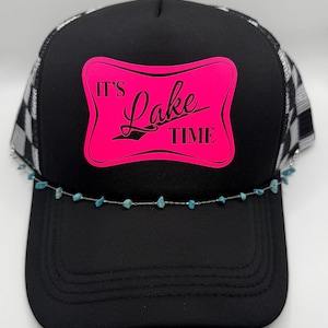 May include: Black trucker hat with a checkered pattern on the mesh and a pink rectangle with the words "It's Lake Time". A silver chain with turquoise beads is attached to the brim.